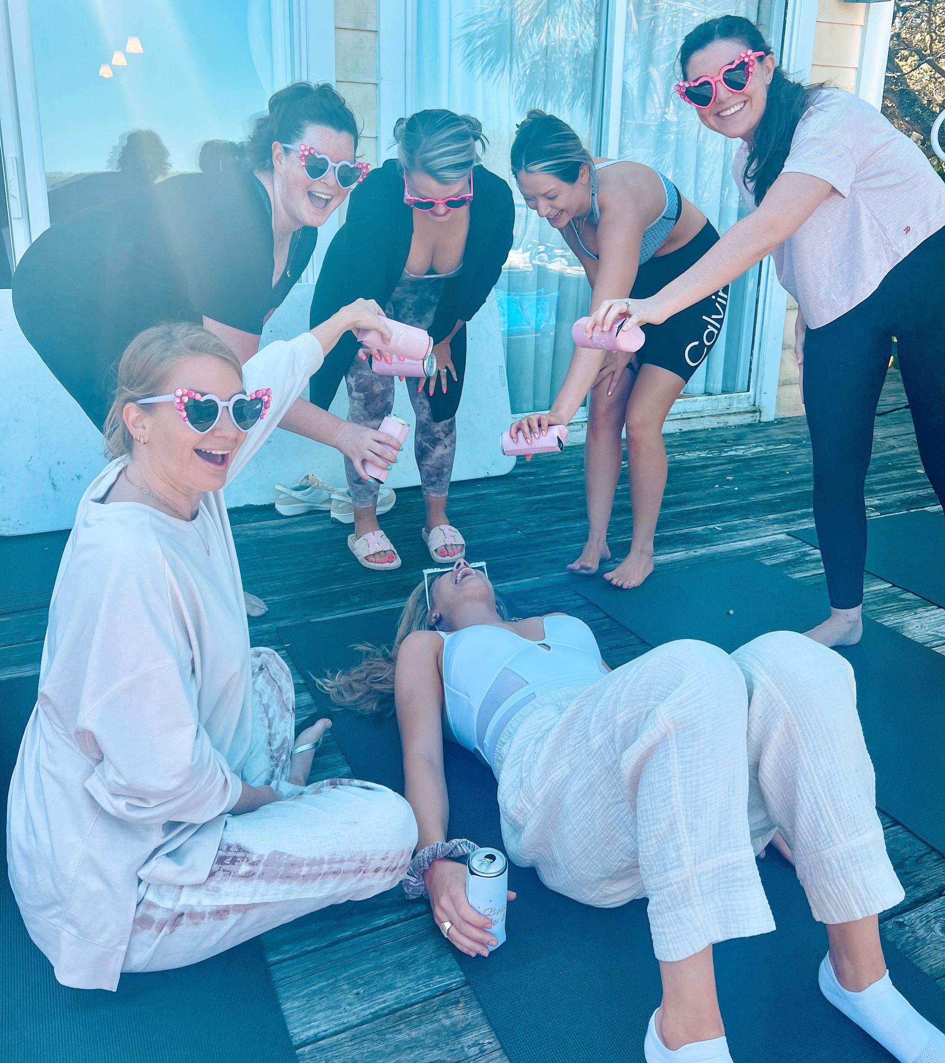 Six women on a sunlit wooden deck with yoga mats, wearing pink heart sunglasses and laughing as they pour canned drinks over a woman lying back.