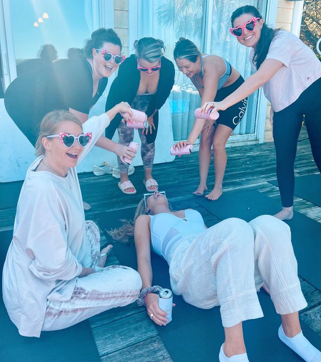 Six women on a sunlit wooden deck with yoga mats, wearing pink heart sunglasses and laughing as they pour canned drinks over a woman lying back.