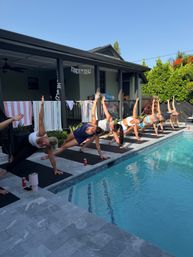 Sunlit backyard poolside yoga — a row of participants on mats holding side-plank poses along a tiled pool beside a covered patio and lush greenery.