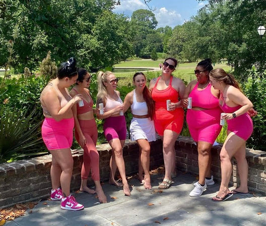 Seven friends in bright pink and white activewear laugh and pose by a low brick wall in a leafy park on a sunny day, each holding a canned drink.