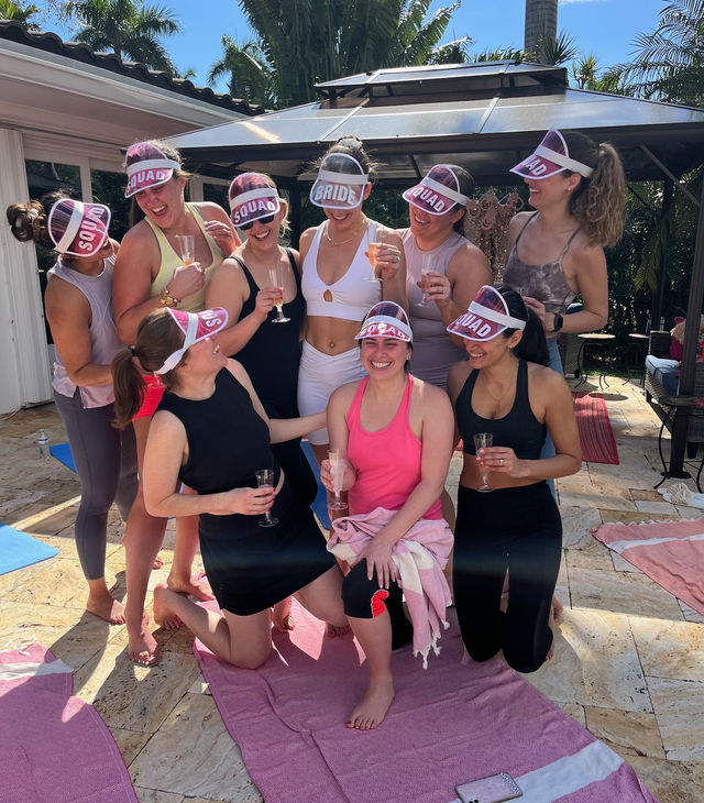 Bachelorette party on a sunny tropical backyard patio with palm trees, bride in white at center and friends wearing pink "SQUAD" visors, laughing and holding champagne flutes on pink towels
