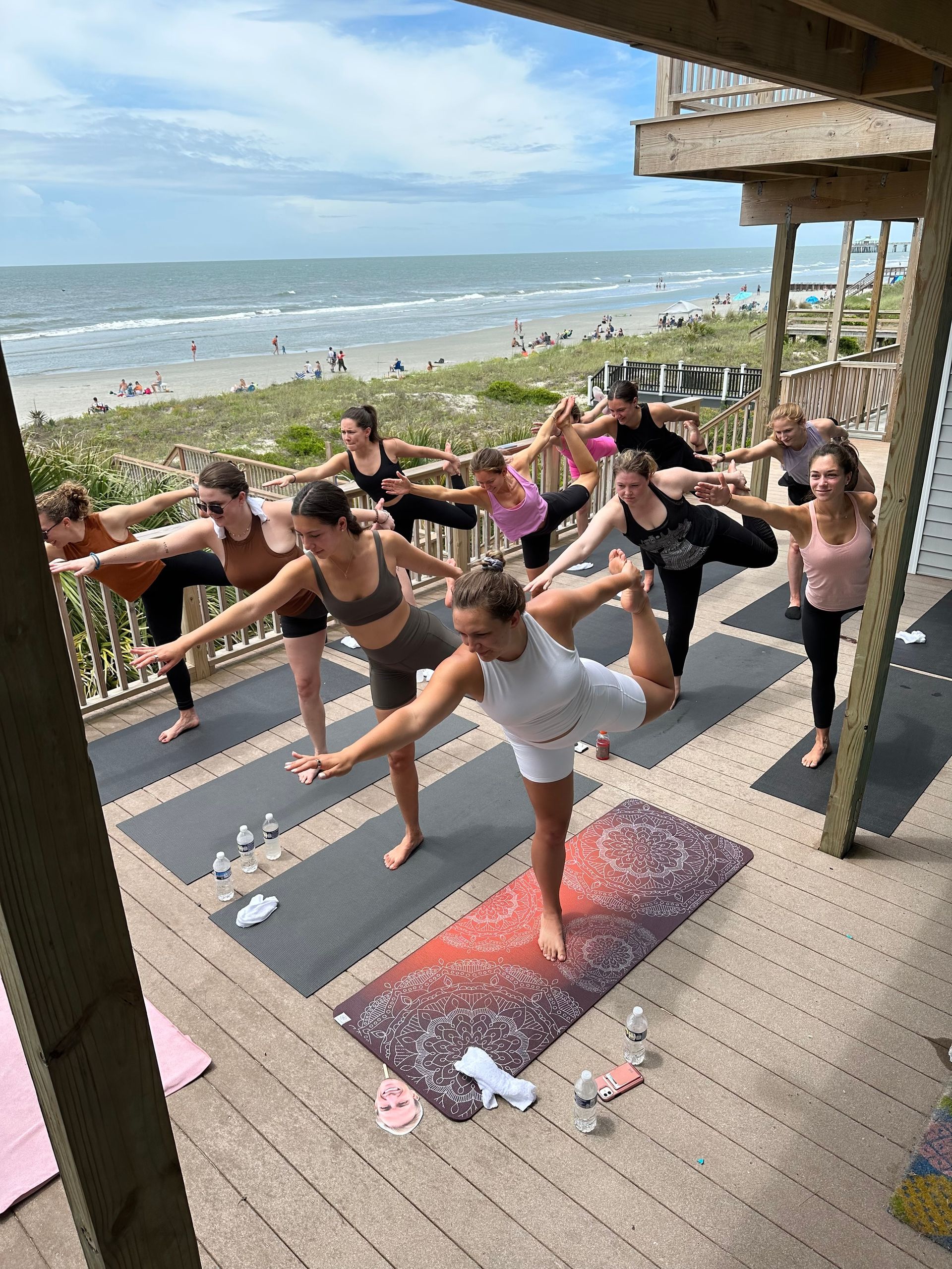 Beachfront group yoga class on a wooden deck — participants holding dancer pose on mats with ocean view and sandy shoreline in the background.