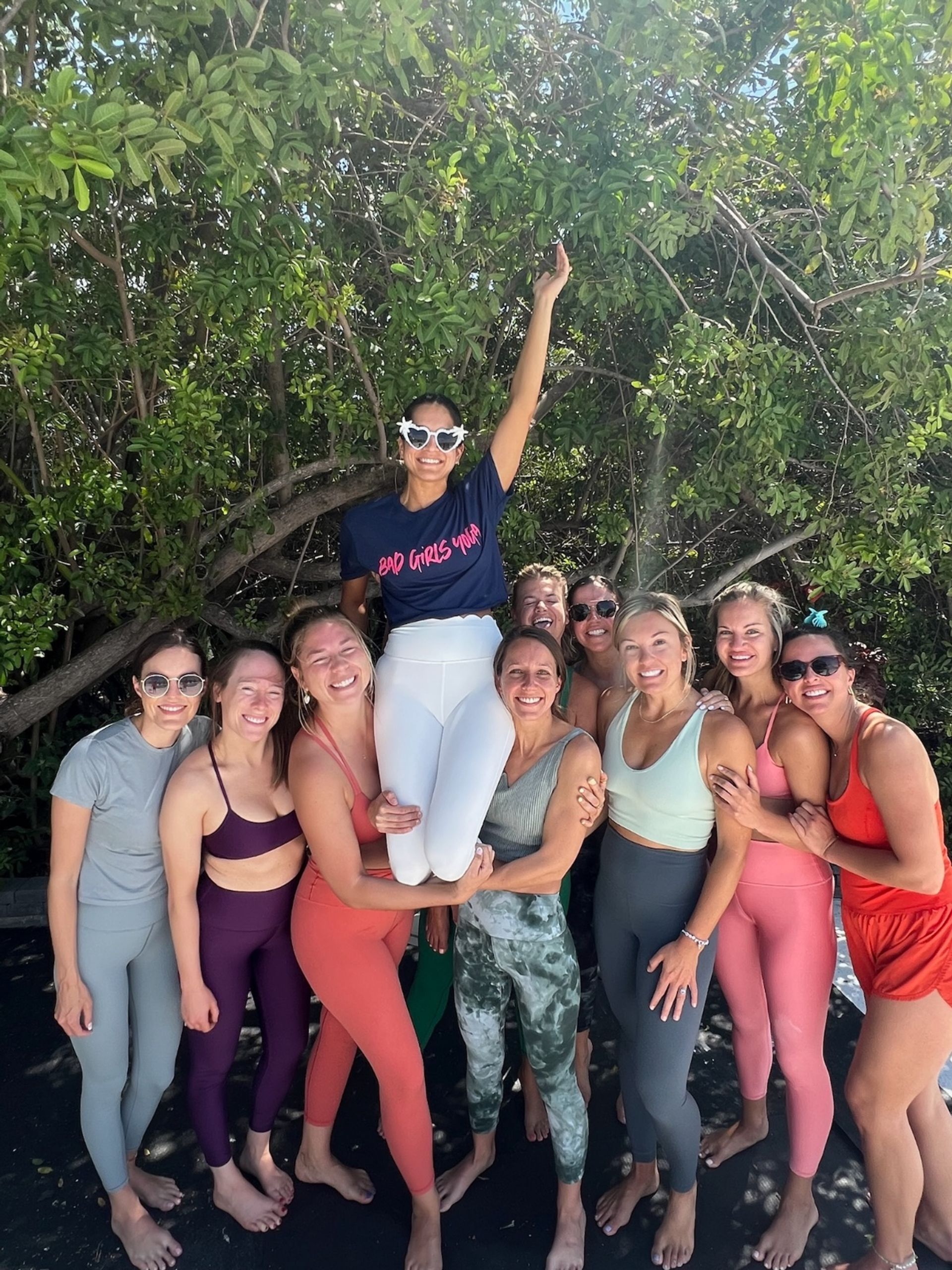 Women’s yoga retreat group photo: smiling women in colorful activewear posing barefoot under tropical mangrove trees on a sunny beach, one woman lifted triumphantly wearing playful sunglasses.