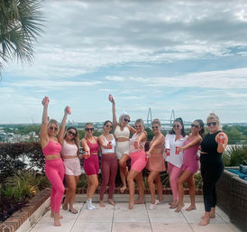 Group of women in pink and white activewear on a rooftop terrace holding canned drinks, posing cheerfully with a waterfront suspension bridge and city skyline in the background.