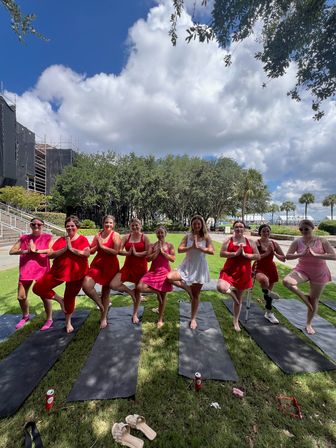 Outdoor group yoga at a waterfront park — nine people in red, pink, and white dresses balancing in tree pose on black mats under puffy clouds, blue sky, and shady trees.