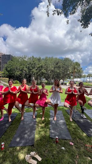 Row of women in red and pink doing tree-pose yoga on black mats in a waterfront park with palm trees, green lawn and dramatic puffy clouds.