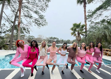 Nine women in coordinated pink and mauve activewear and heart-shaped sunglasses striking a one-leg pose on yoga mats by a pool at a lakeside backyard with palm trees.