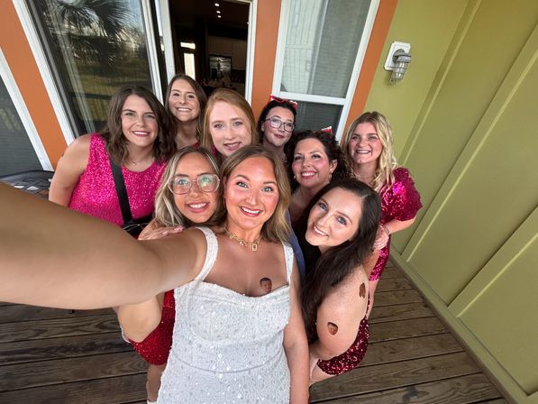 Happy bridal party group selfie on a wooden porch with colorful siding, women in sequined red dresses and one in a white dress, smiling close-up faces.