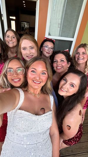 Cheery group selfie on a wooden porch — bride in a white dress surrounded by smiling friends in pink sequined dresses, festive bridal party vibe in front of a green house exterior and windows.