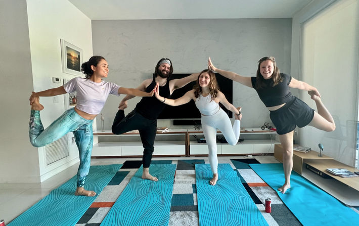 Four friends smiling and balancing in dancer yoga pose on blue mats in a bright modern living room, touching palms in an at-home yoga session.