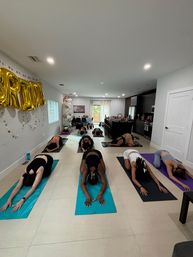 Group yoga class practicing child’s pose on mats in a bright open-plan living room, with gold celebration balloons and a modern kitchen in the background