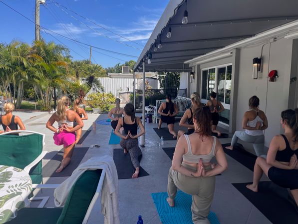 Sunny outdoor patio yoga class with women on mats in low lunge and reverse-prayer hands under an awning beside palm trees and tropical landscaping.