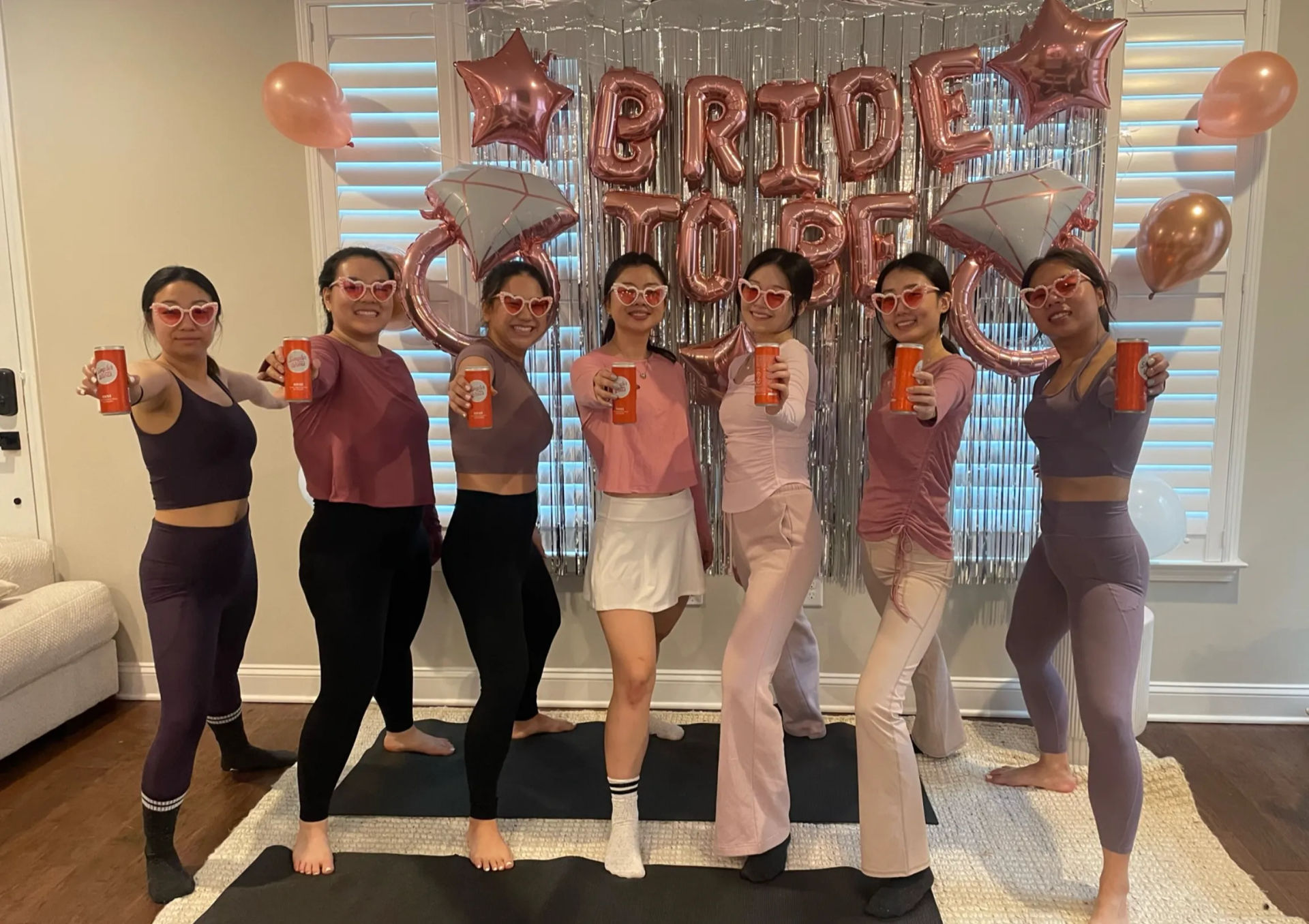 Seven women in pink and mauve activewear wearing heart-shaped sunglasses pose in a living room bachelorette party, holding orange canned drinks in front of a rose-gold "BRIDE TO BE" balloon backdrop with star and ring balloons.