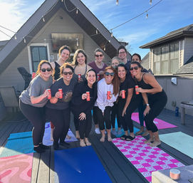 Smiling group of women on a sunny rooftop deck with yoga mats and string lights, holding canned drinks and posing together.
