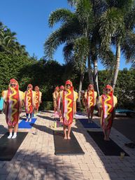 Adults in hot dog costumes standing on yoga mats for a sunny poolside yoga class beneath palm trees