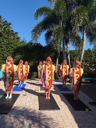 Adults in hot dog costumes standing on yoga mats for a sunny poolside yoga class beneath palm trees