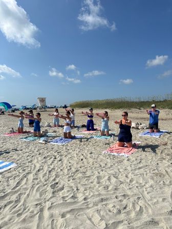 Group beach yoga class kneeling on towels on a sunny sandy shore, stretching arms under a blue sky with dunes and a lifeguard stand nearby.