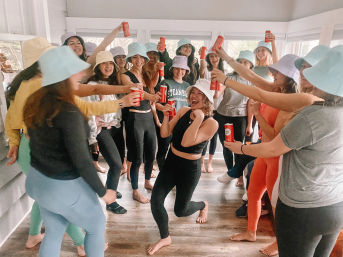 Large group of friends in colorful bucket hats and activewear cheering indoors, barefoot and raising red drink cans while a laughing woman in black leggings poses center on a wood floor in a bright sunroom-style room.