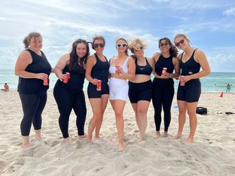 Seven friends in black and white swimwear holding red cans, smiling and posing barefoot on a sandy ocean beach with blue sky and waves