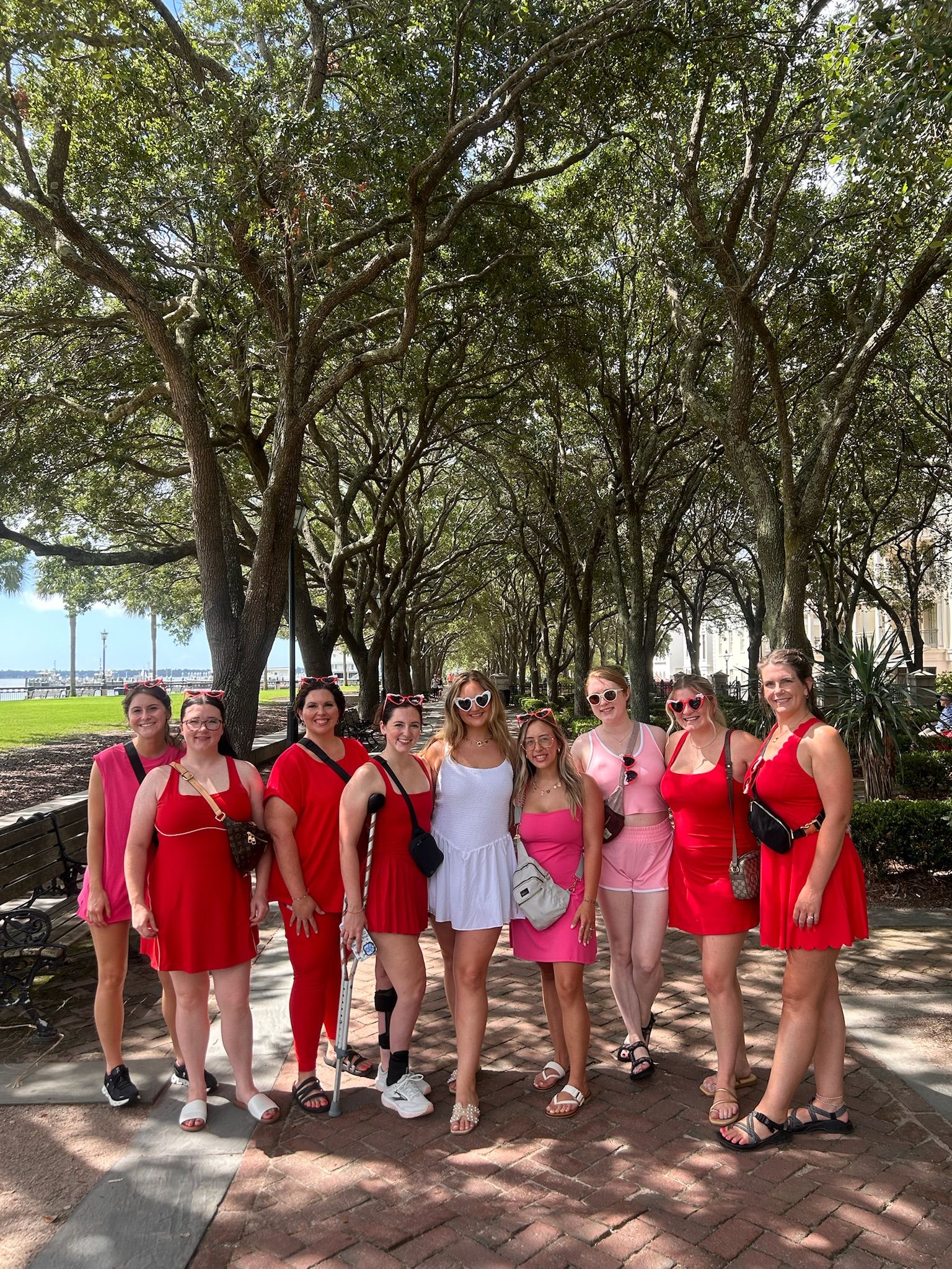 Group photo of women in red, pink and white summer dresses posing on a brick waterfront promenade under an arching oak tree canopy on a sunny day