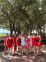 Group photo of women in red, pink and white summer dresses posing on a brick waterfront promenade under an arching oak tree canopy on a sunny day