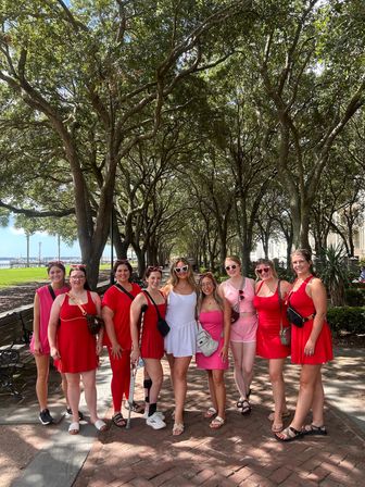 Group photo of women in red, pink and white summer dresses posing on a brick waterfront promenade under an arching oak tree canopy on a sunny day