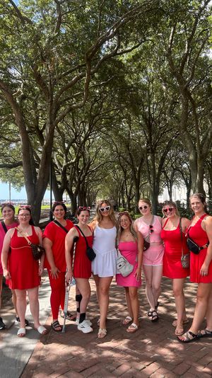 Ten women in red, pink and white outfits smiling and posing together under a shaded canopy of oak trees on a waterfront park promenade with benches and a distant marina.