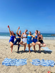 Six women in blue and white athletic swimwear balancing on one leg and holding red cans on a sunny sandy beach, striped blue-and-white towels on the sand with ocean waves and a clear blue sky in the background.
