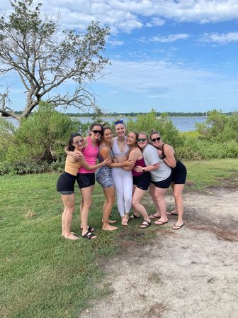 Seven women in casual activewear hugging and smiling on a grassy lakeside path under a blue summer sky