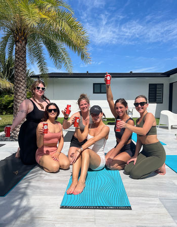 Six women in athletic wear on blue yoga mats on a sunny backyard patio under a palm tree, smiling and cheering while toasting with red canned drinks against a bright blue sky.