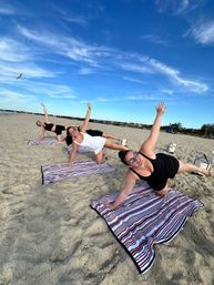 Three women in athletic wear doing side-plank exercises on striped towels on a sunny sandy beach under a bright blue sky with a seagull overhead.