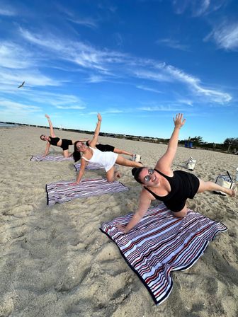 Three women in athletic wear doing side-plank exercises on striped towels on a sunny sandy beach under a bright blue sky with a seagull overhead.