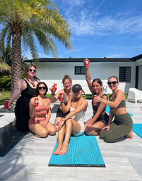 Six women in swimwear and activewear on yoga mats at a tropical poolside patio, smiling and toasting with red canned drinks under a palm tree and bright blue sky.