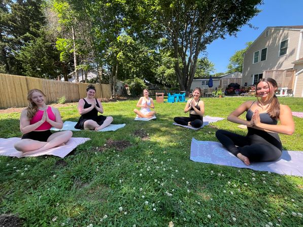 Five women smiling in seated yoga pose on mats during a sunny suburban backyard yoga session on a green lawn with large shade trees, a wooden fence and house in the background.