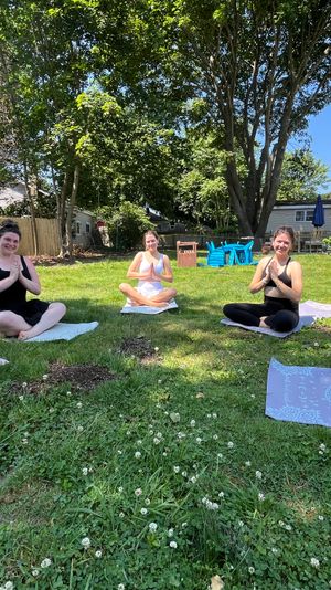 Five women smiling in seated yoga pose on mats during a sunny suburban backyard yoga session on a green lawn with large shade trees, a wooden fence and house in the background.