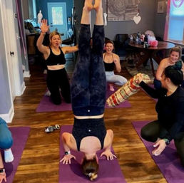 Lively indoor yoga class on purple mats with a woman holding a headstand as classmates cheer and spot her on a hardwood floor