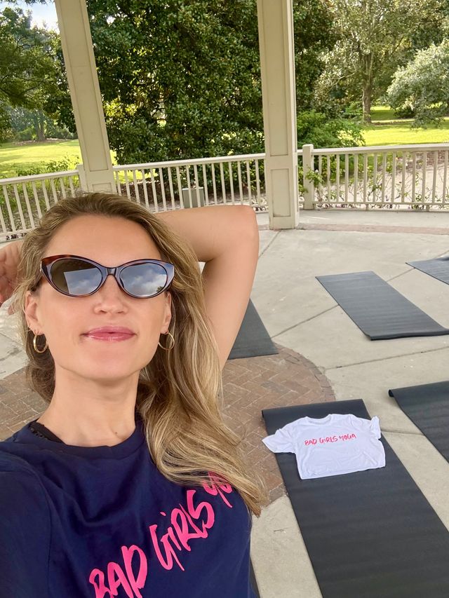 Woman in cat-eye sunglasses taking a selfie in a covered park gazebo set up for outdoor yoga — black mats on concrete, navy tee with pink script and a white tee with pink lettering on one mat, green trees beyond.