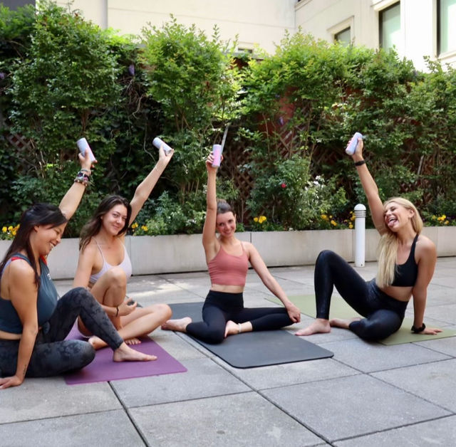 Four women in workout clothes on yoga mats in a city courtyard, raising water bottles and laughing after an outdoor yoga/stretch session by green hedges.