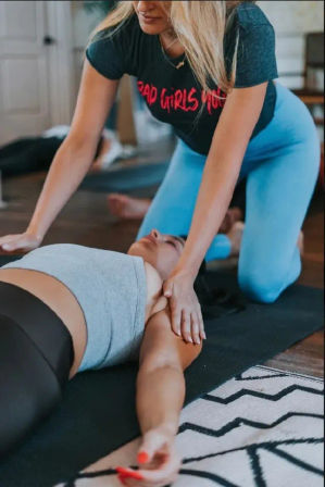 Hands-on yoga stretch — instructor kneels over a student guiding a shoulder and chest stretch on a mat in an indoor fitness class.