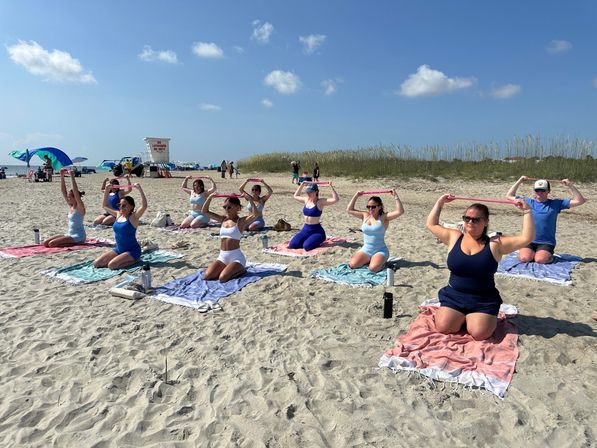 Sunny beach workout class on a sandy shore — participants kneeling on towels and holding pink resistance bands overhead, lifeguard tower and dunes under a blue sky