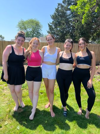 Five smiling women posed arm‑in‑arm in a sunny suburban backyard wearing sporty tops, shorts and leggings in front of a wooden fence and trees