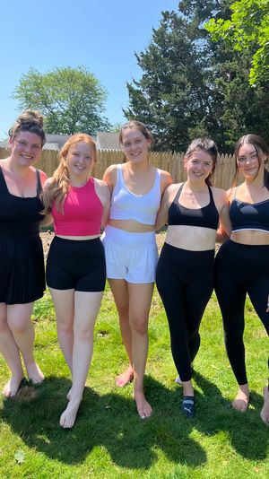 Five smiling women posed arm‑in‑arm in a sunny suburban backyard wearing sporty tops, shorts and leggings in front of a wooden fence and trees