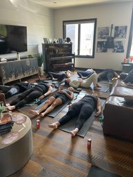 Sunlit home yoga class with several people lying in Savasana on mats in a cozy living room with hardwood floors, an upright piano, wall-mounted guitar photos and a large window.