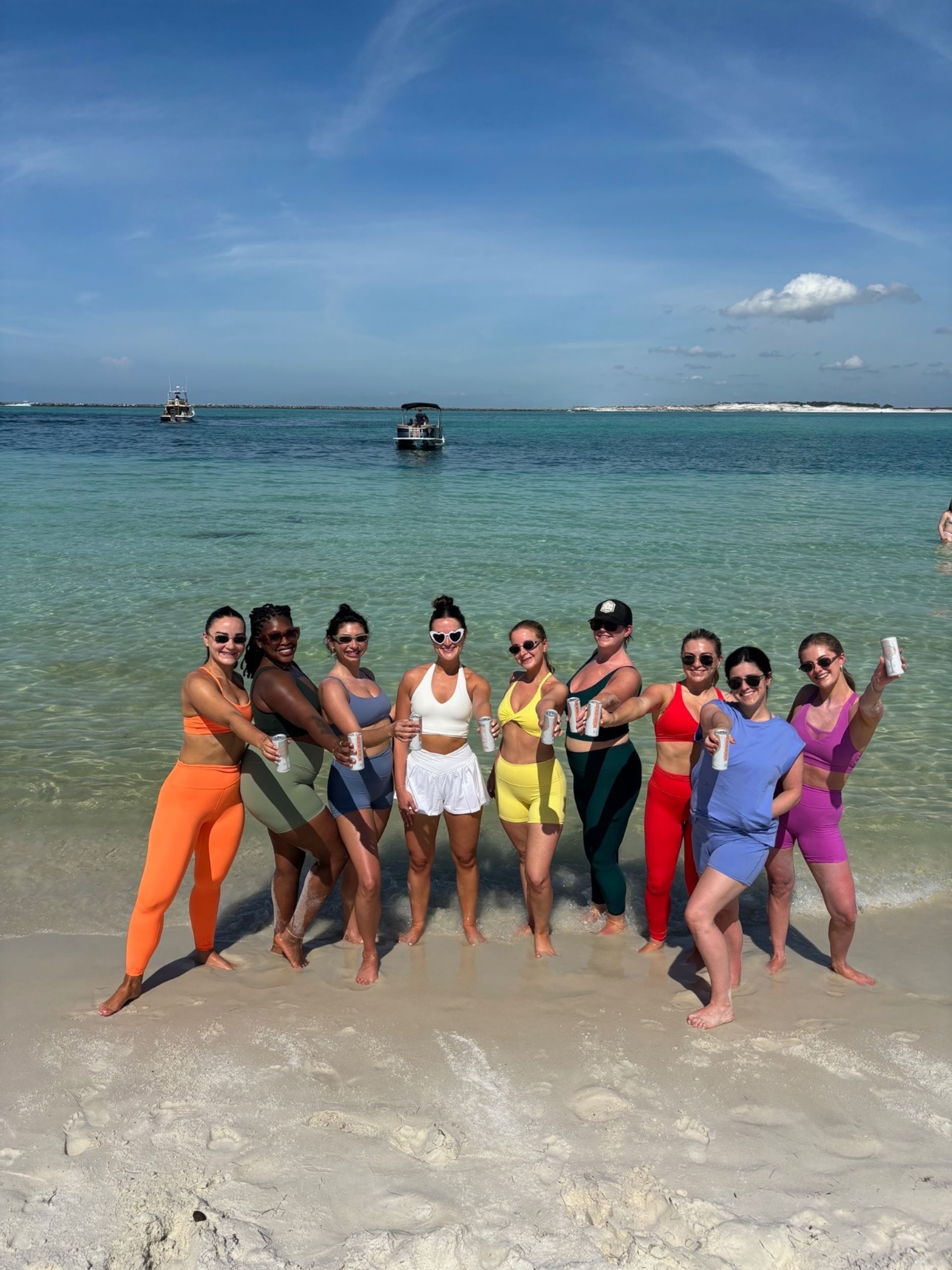 Nine women in colorful swimwear and activewear standing in shallow turquoise surf, smiling and toasting with canned drinks on a sunny beach with small boats and a clear blue sky in the background.