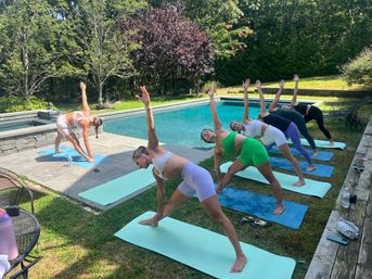 Sunlit poolside yoga session with six women on colorful mats doing triangle poses beside a backyard swimming pool surrounded by trees and lawn.