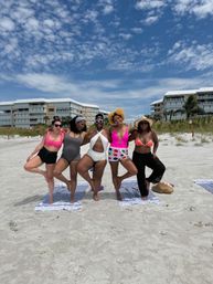 Five friends in colorful swimsuits linking arms and posing on a sandy beach with beachfront condos and a bright blue, partly cloudy sky.