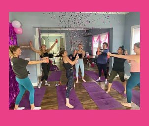 Women celebrating on purple yoga mats in a bright living-room-style studio, popping confetti and raising drink cans amid pink balloons and streamers.
