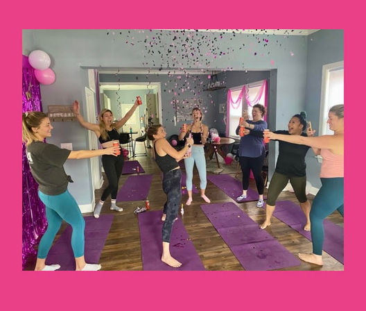 Women celebrating on purple yoga mats in a bright living-room-style studio, popping confetti and raising drink cans amid pink balloons and streamers.