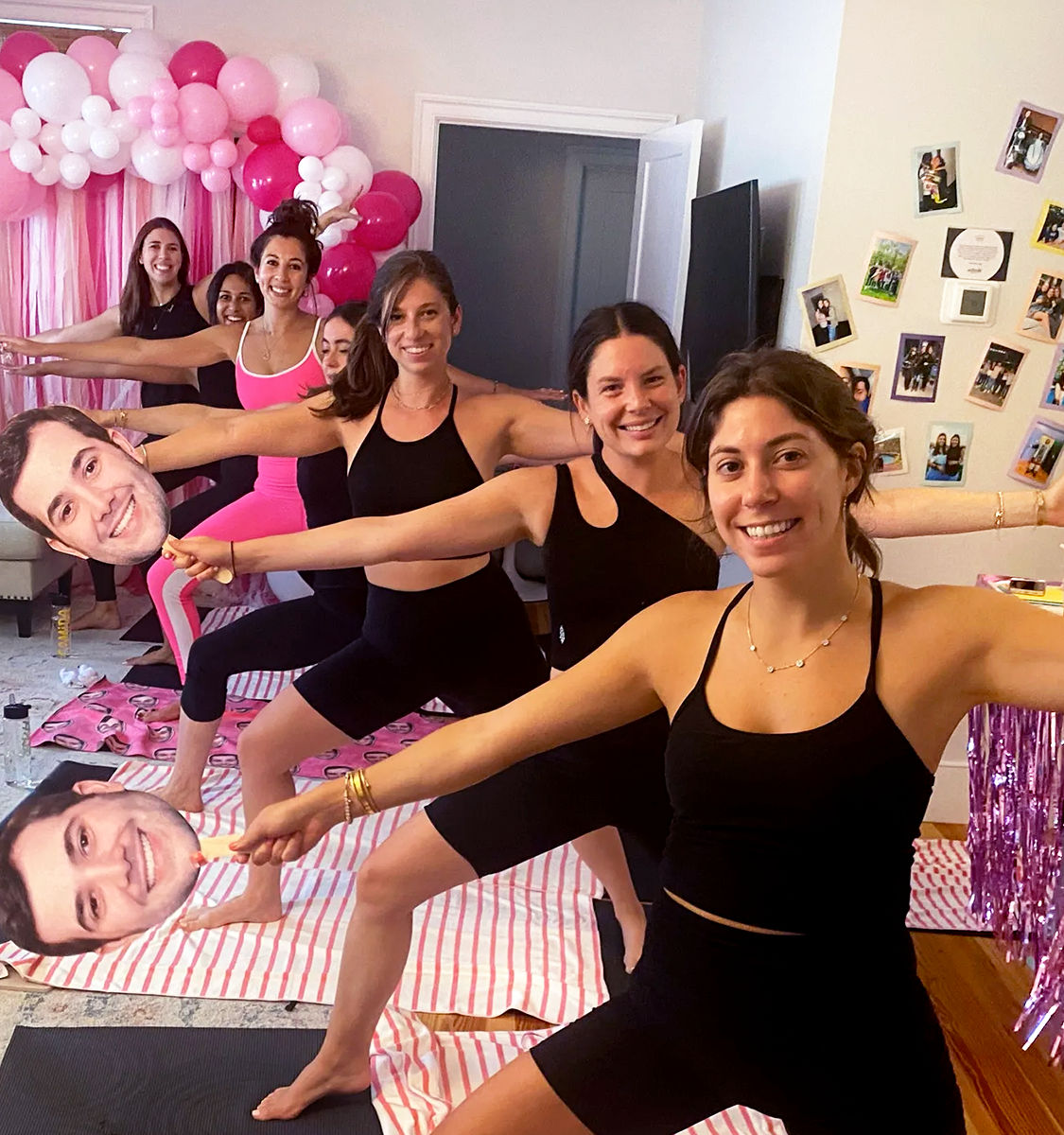 Six women in a living room doing warrior yoga poses on striped mats at an at-home pink-themed party, smiling and holding oversized cutout faces under a pink-and-white balloon arch.