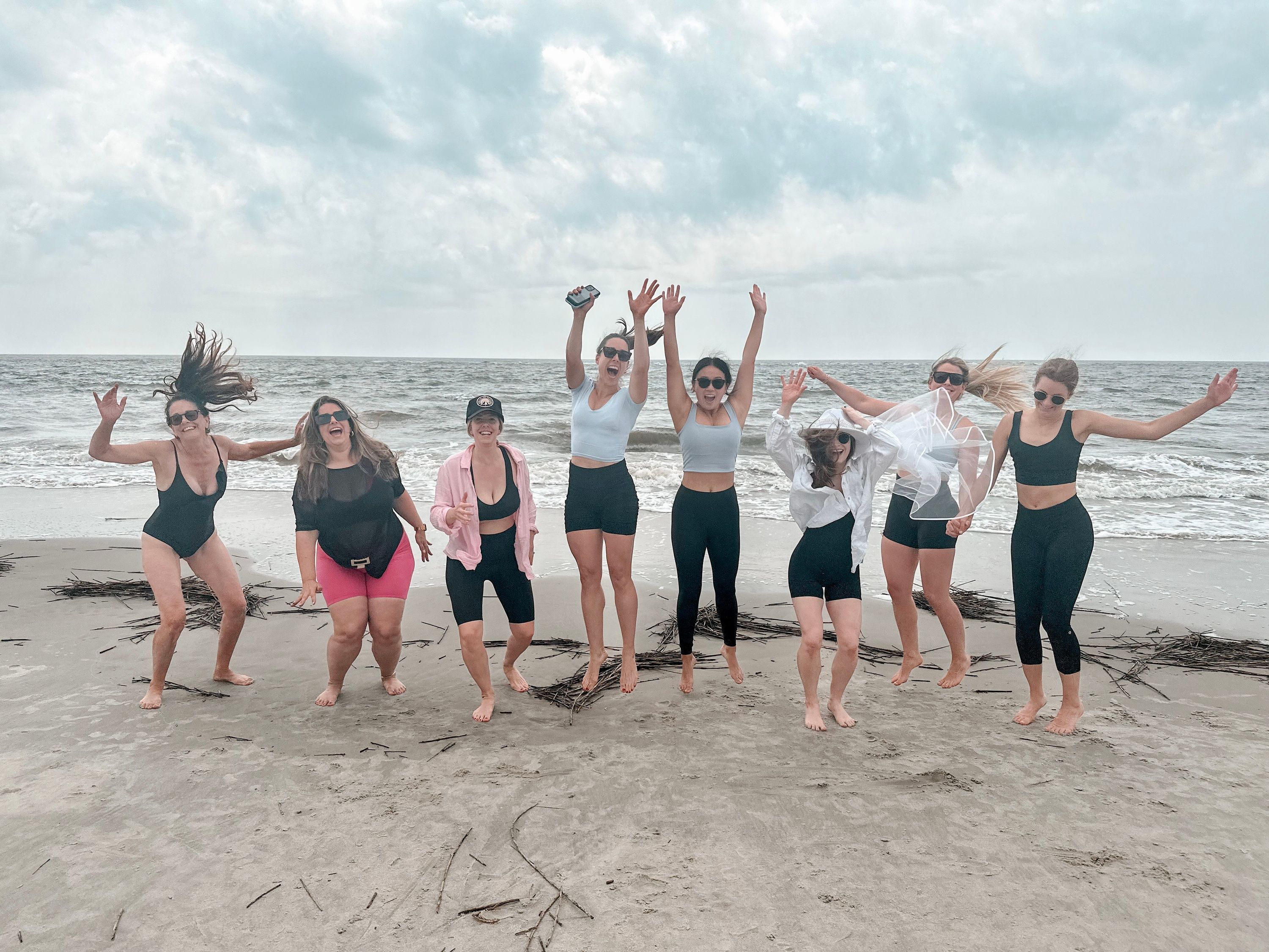 Eight women friends jumping and cheering barefoot on a sandy ocean beach with waves and an overcast sky, wearing swimsuits and activewear — lively seaside group photo.